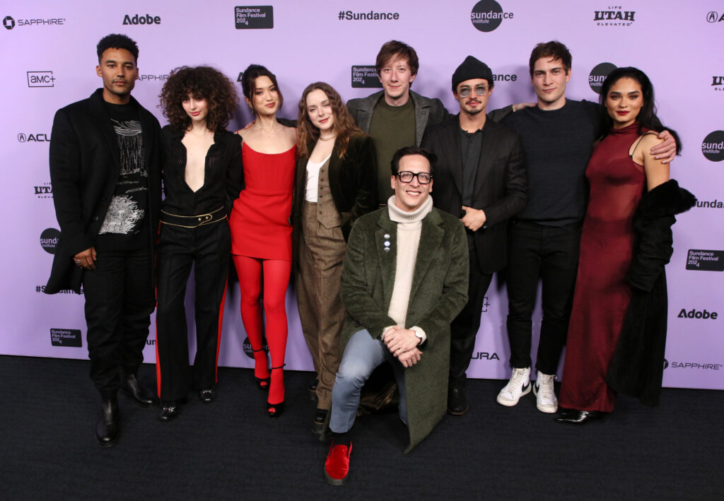 Greg Jardin smiles as he kneels in front of Devon Terrell, Nina Bloomgarden, Reina Hardesty, Madison Davenport, David Thompson, Gavin Leatherwood, James Morosini, and Brittany O’Grady. They are posing in front of a white 2024 Sundance Film Festival backdrop.
