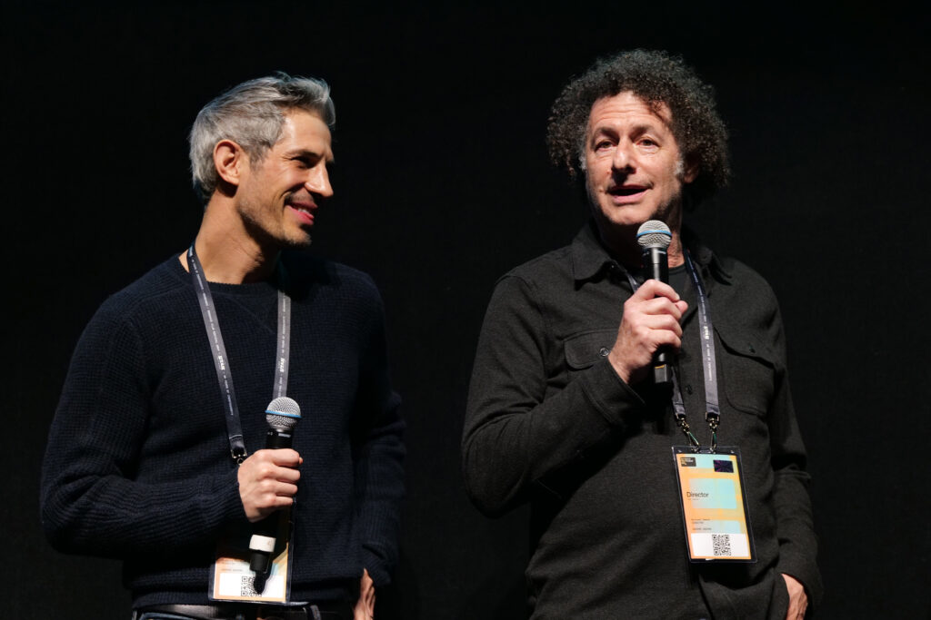 Gregory Kershaw and Michael Dweck stand in front of a black backdrop. Both are holding microphones.